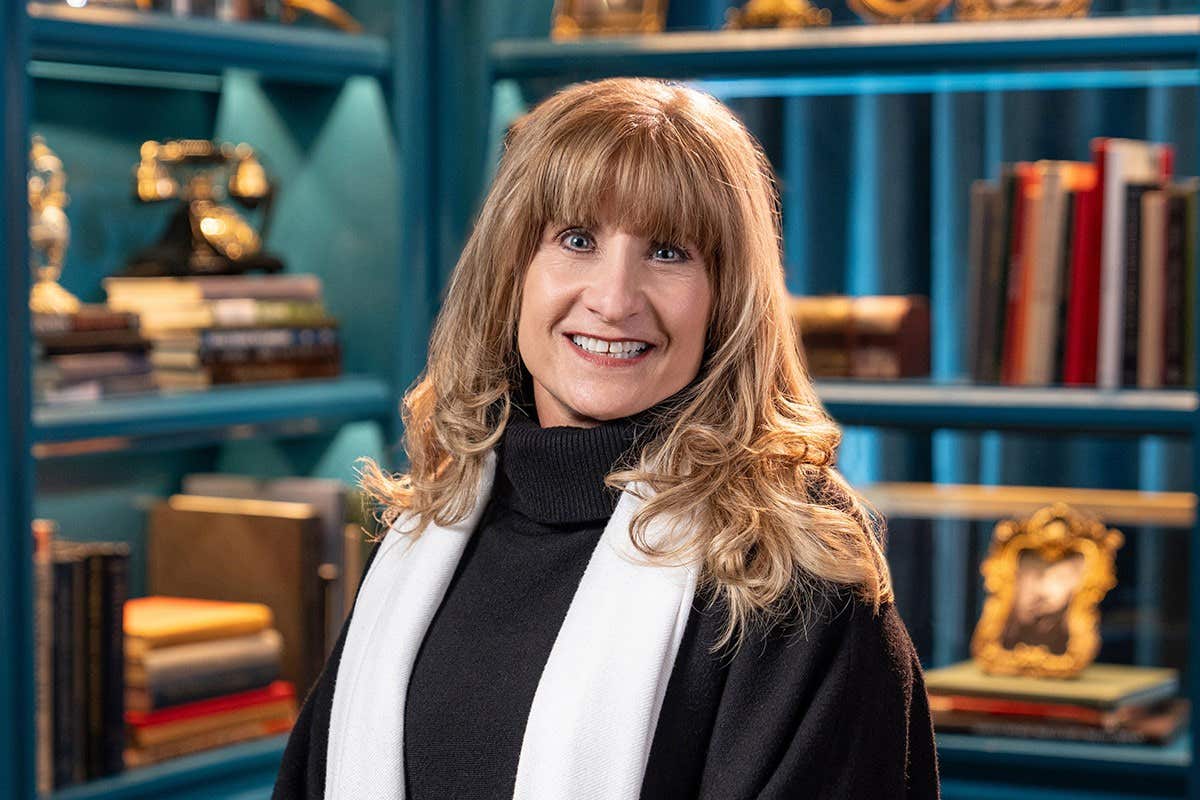 A woman with long, blonde hair wearing a black sweater and white scarf stands smiling in a library.