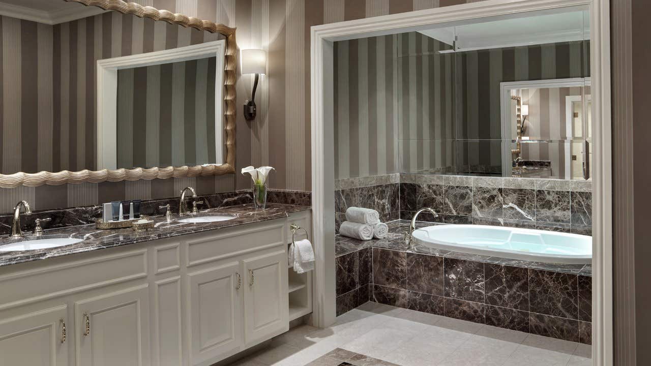 Luxurious bathroom with two sinks, large mirror, marble countertop, and a bathtub surrounded by dark marble tiles.