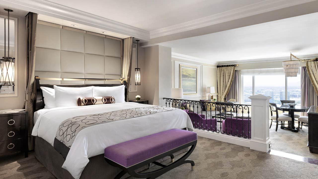 Elegant hotel room with a king-size bed, seating area, and large window offering a city view in the background.