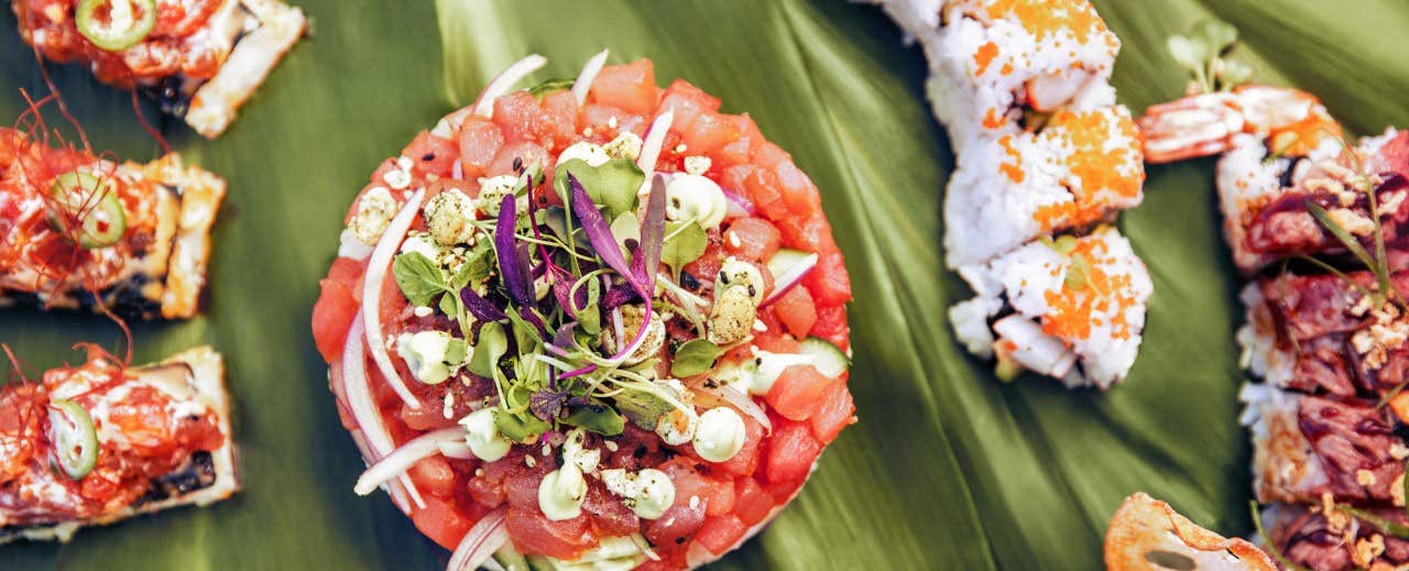 A close-up of assorted sushi and a dish of diced seafood garnished with greens on a banana leaf background.