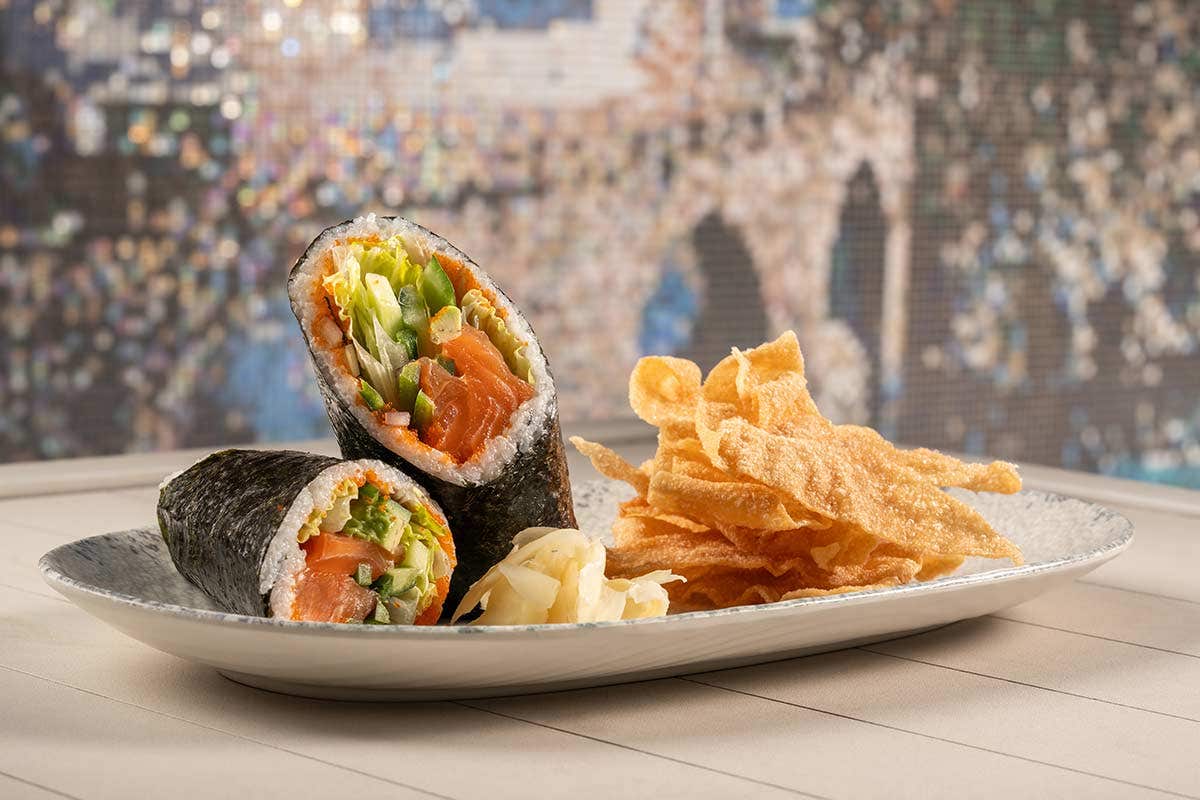 Two sushi hand rolls with raw salmon and vegetables, served with crispy wonton chips and pickled ginger on a white plate.