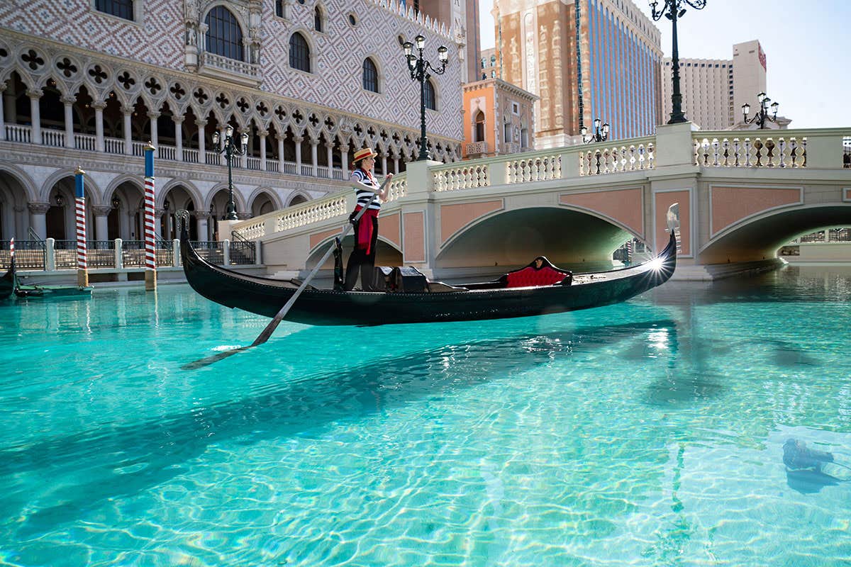 A gondolier rowing a gondola in a turquoise canal, surrounded by Venetian-style architecture and bridges.