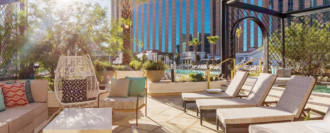 Outdoor patio area with modern lounge chairs, comfy pillows, and plants, overlooking a sunlit hotel pool and tall buildings.