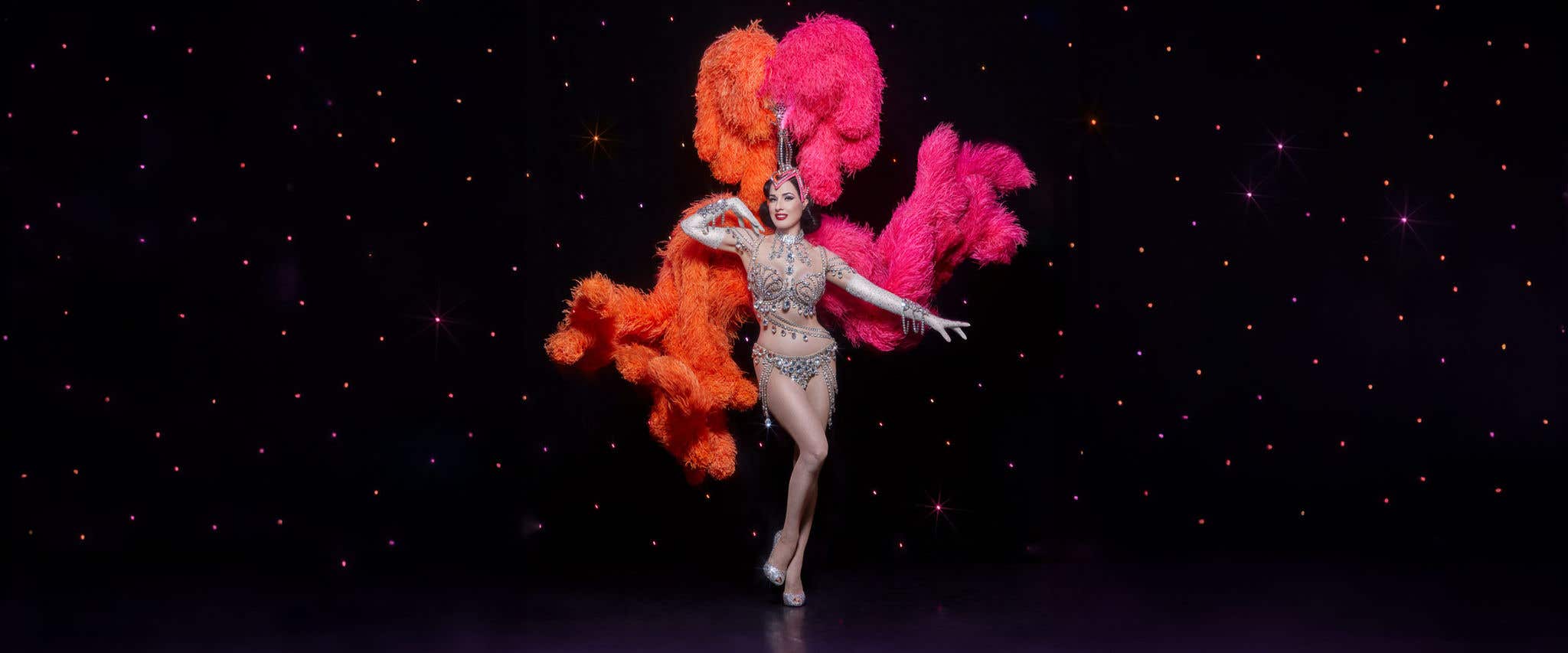 Dita Von Teese in a diamond-covered outfit with an orange and pink headdress in front of a starry backdrop