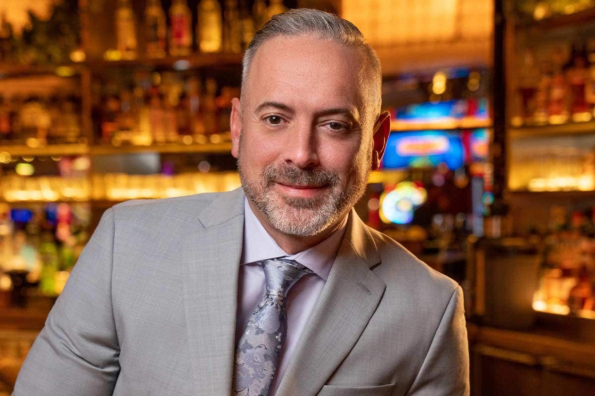 A man in a light grey suit with a tie smiles in front of a warmly lit bar background.