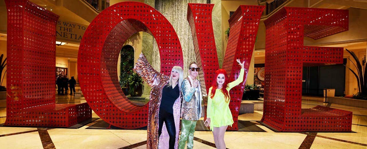 Three people in colorful outfits pose in front of a large red 'LOVE' sculpture inside a spacious building.