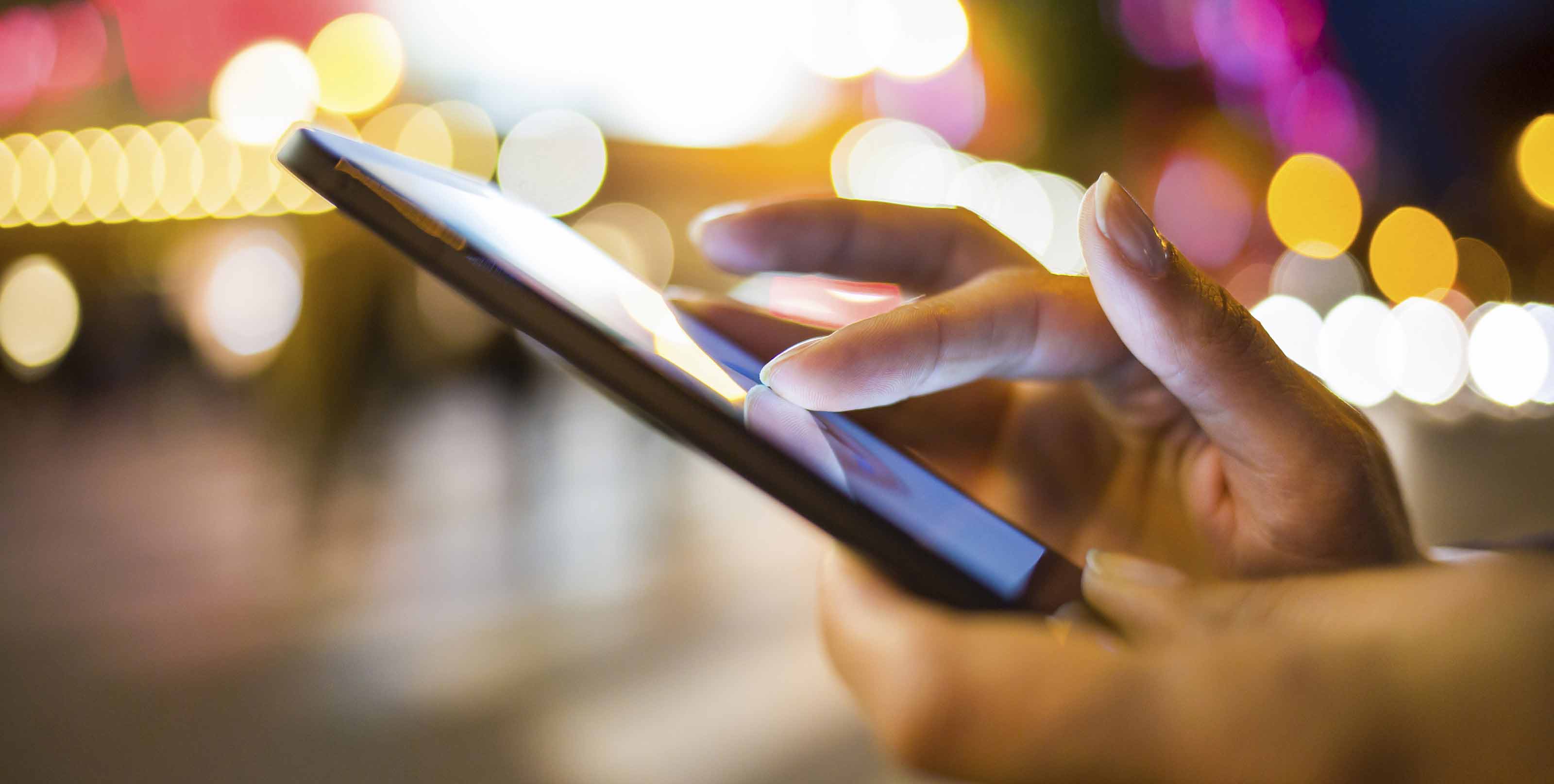 Close-up of a person's hands using a smartphone with a colorful, blurred cityscape in the background.