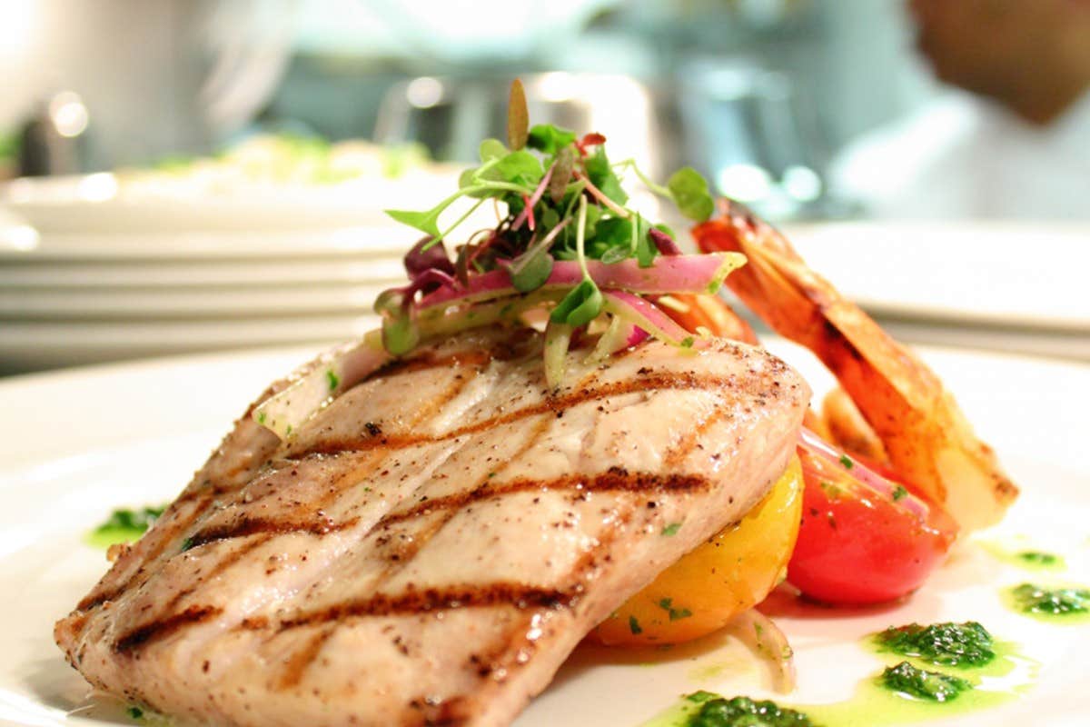 A close-up of a grilled fish fillet topped with microgreens and served with colorful vegetables and a side of sauce.