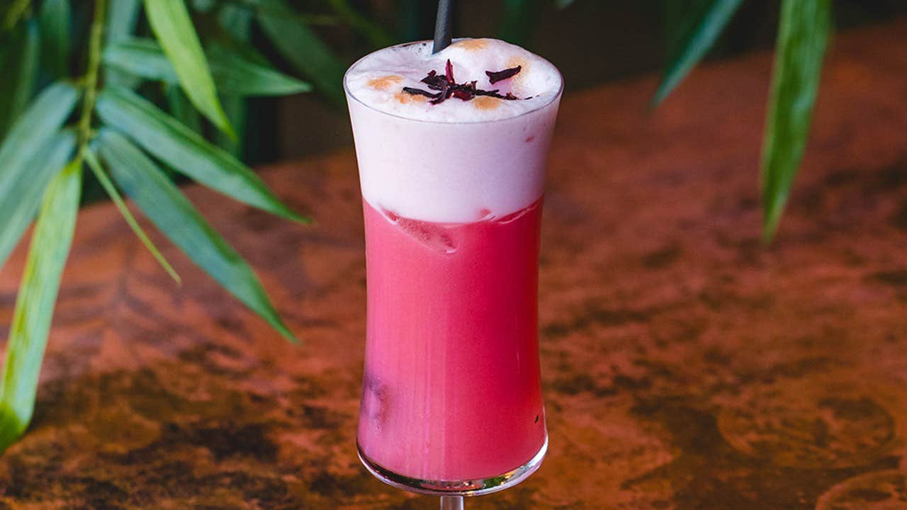 A tall pink drink with a frothy top, garnished with flower petals, on a wooden table with green leaves in the background.