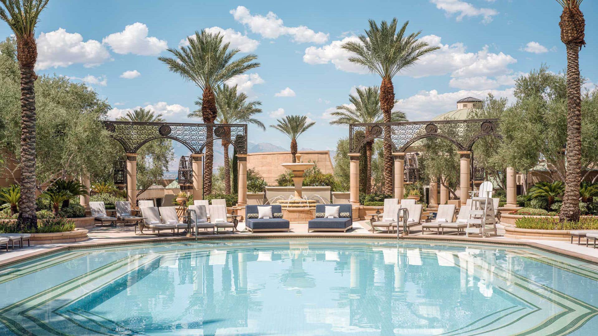 A luxurious swimming pool with lounge chairs, palm trees, and a lavish resort building in the background on a sunny day.