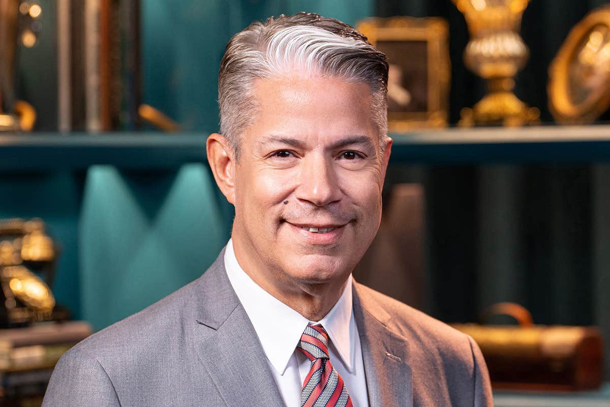 A man in a gray suit and red-striped tie smiles in front of a bookshelf with decorative items.