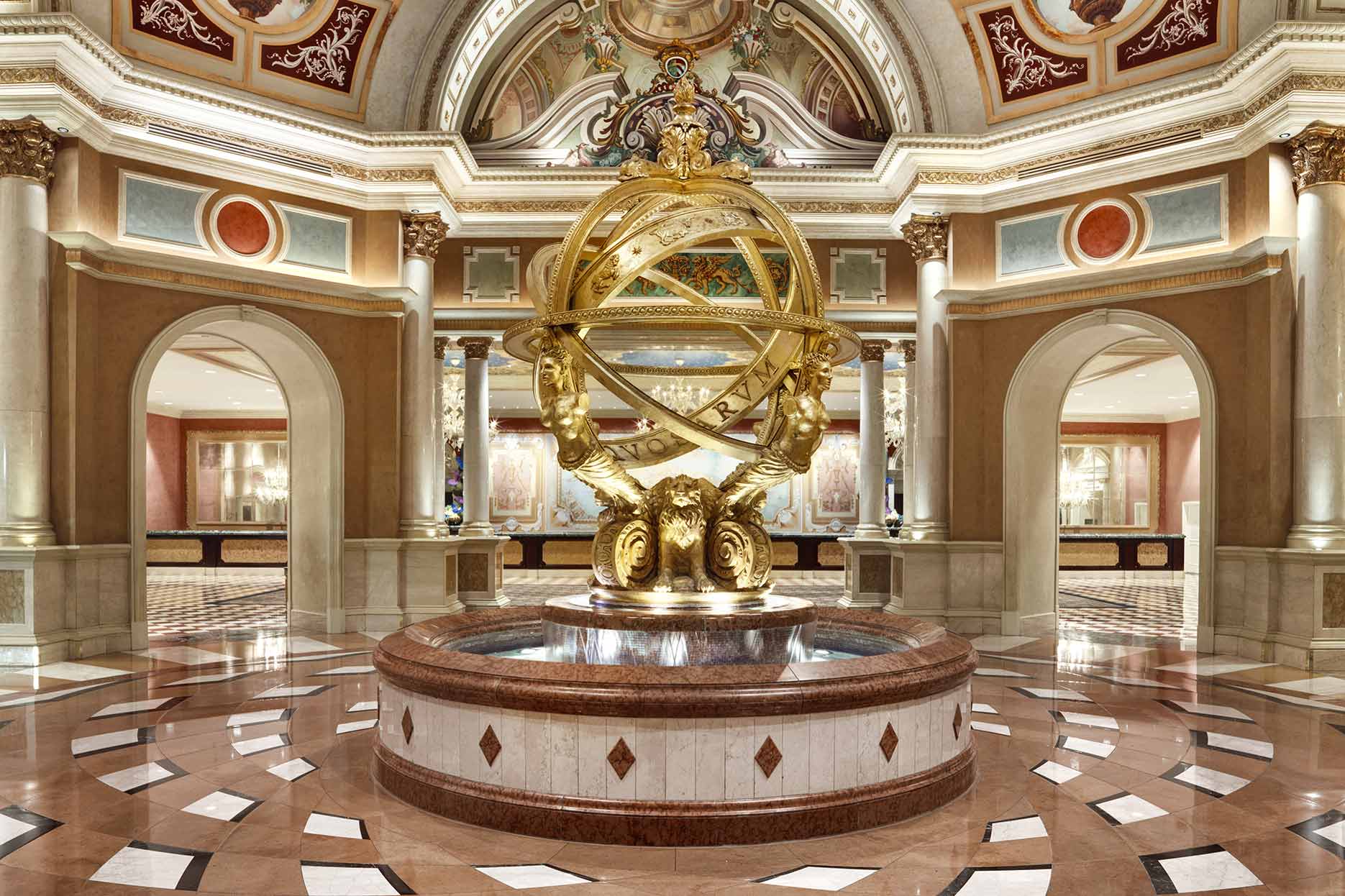 Luxurious hotel lobby with ornate golden globe statue, marble floors, and arched doorways featuring opulent decor.