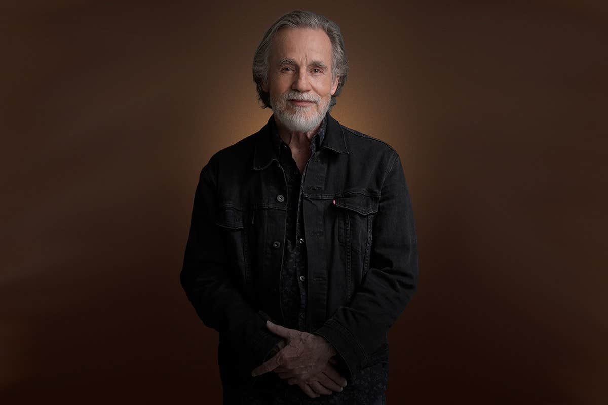 Elderly man with gray hair and beard wearing a black jacket, standing with a calm expression against a brown background.