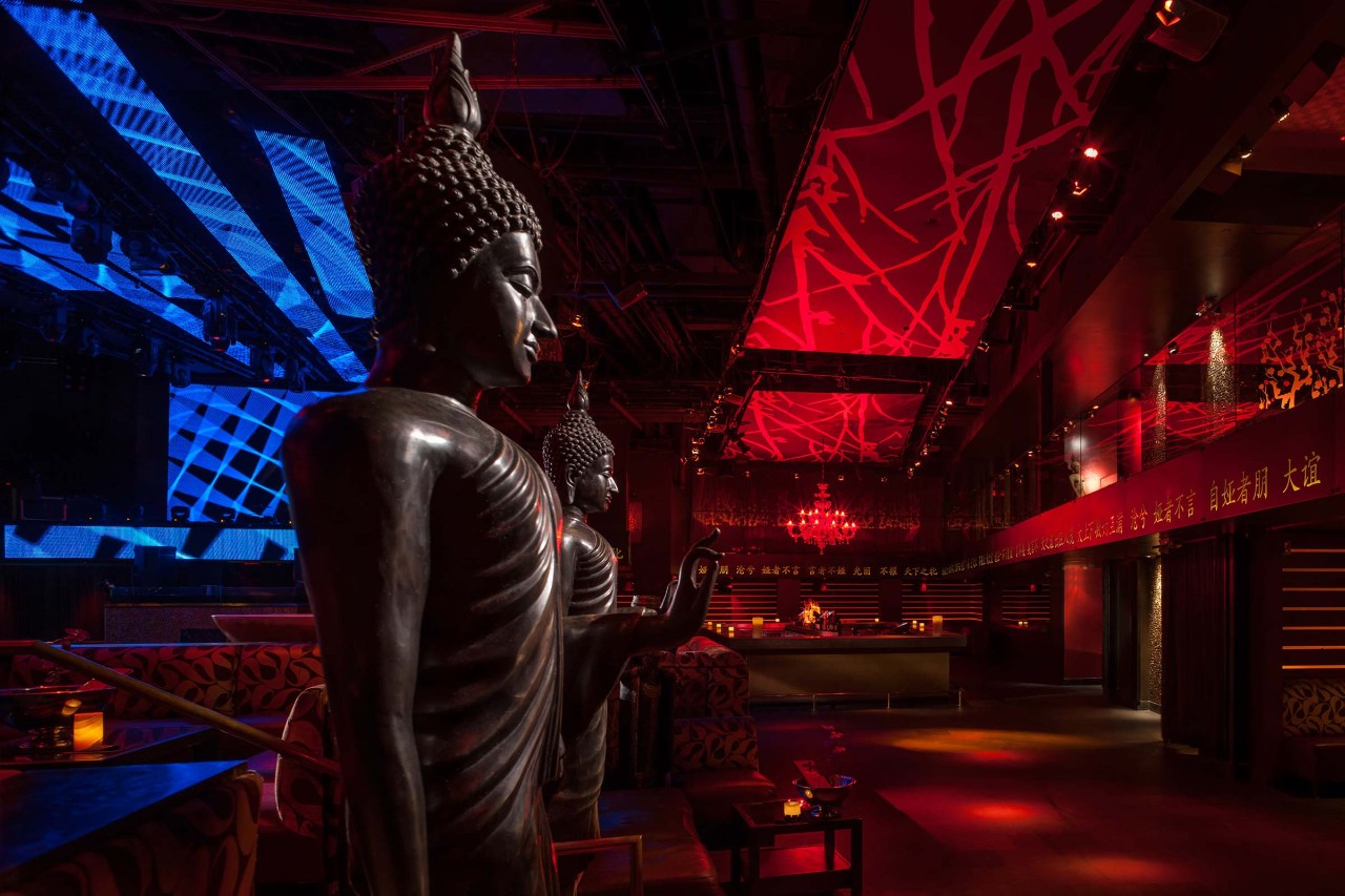 Dimly lit club interior with Buddha statues, red and blue lighting, and abstract ceiling patterns.