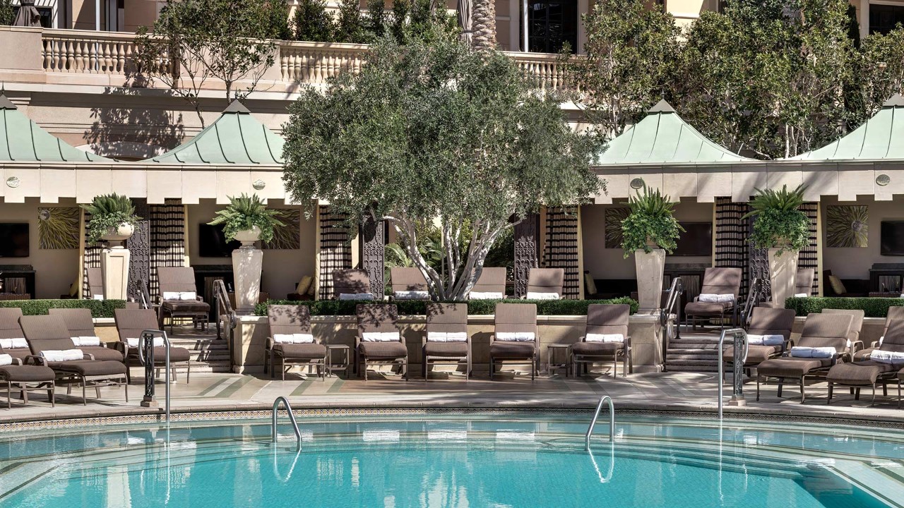 A luxurious hotel pool area with lounge chairs, cabanas, potted plants, and an olive tree in the center.