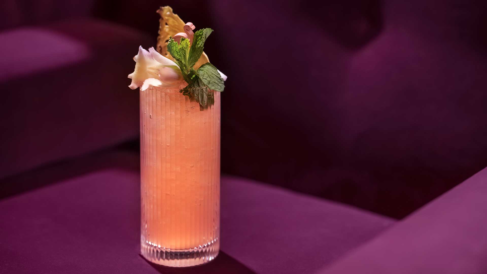 A pink cocktail garnished with mint leaves and a dried fruit slice in a textured glass on a purple surface.