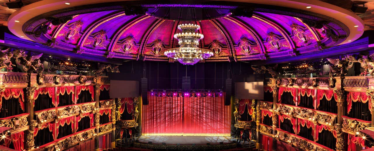 Ornate theater interior with red seats, gold accents, a chandelier, and purple lighting above the stage.