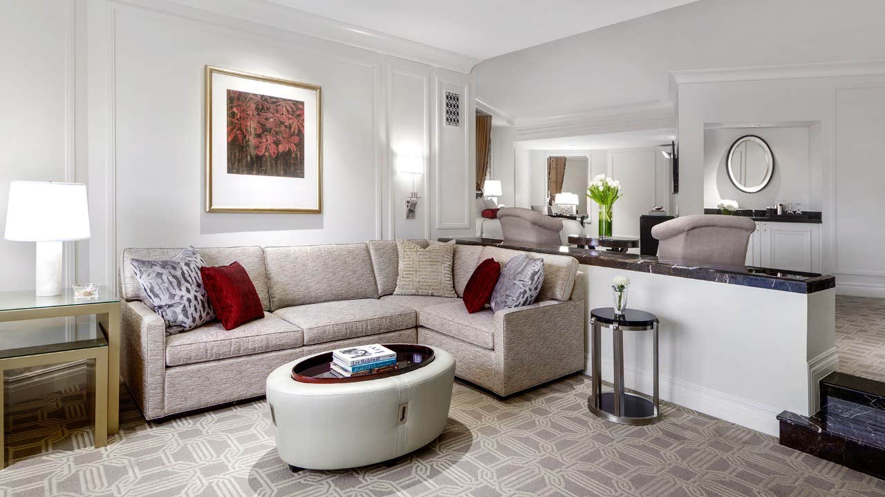 Elegant hotel suite with beige sectional sofa, red pillows, round ottoman, artwork, marble bar counter, and modern décor.