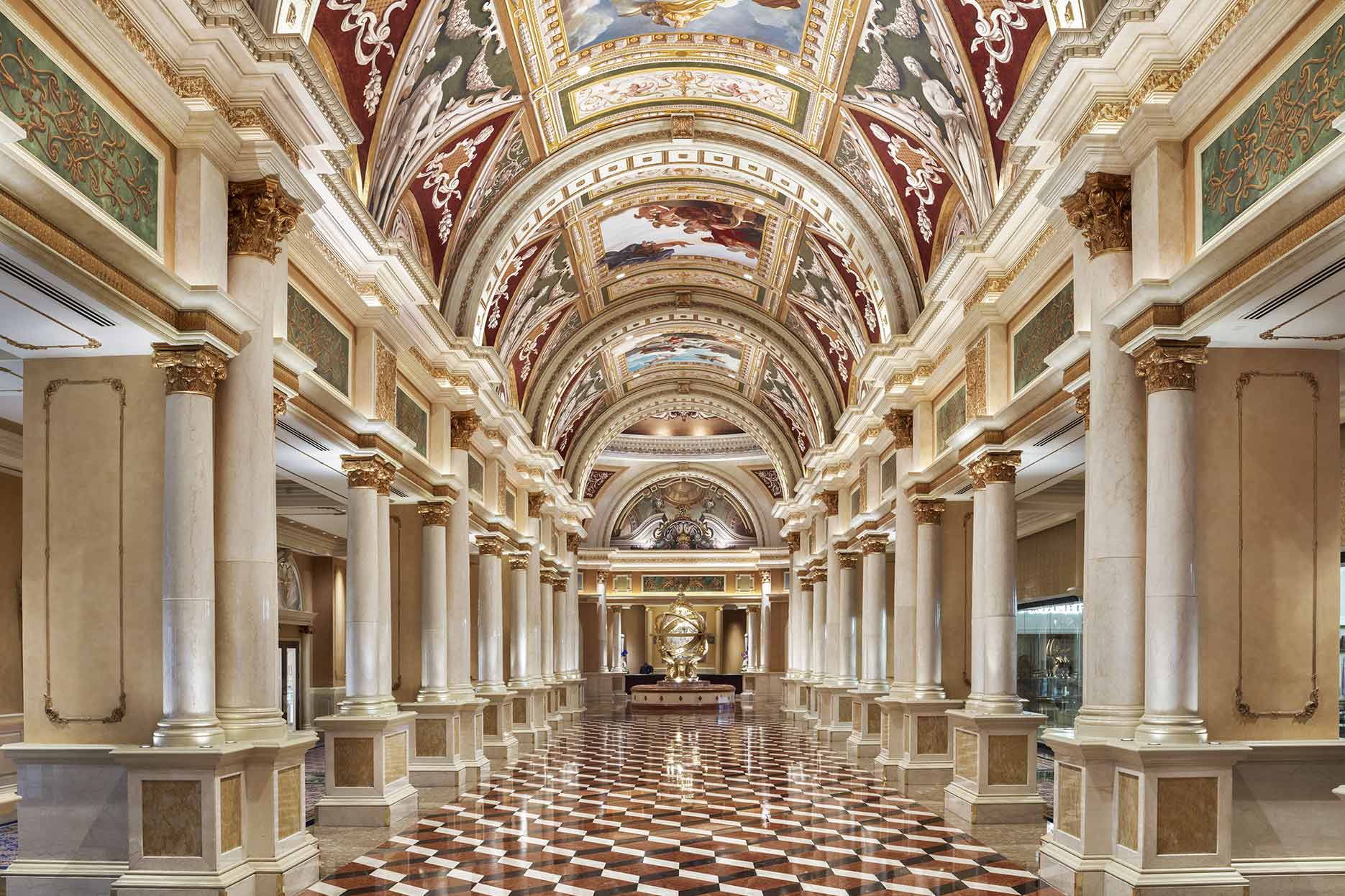 Ornate hallway with grand arches, decorated ceilings, marble columns, and a checkered floor, reminiscent of a palace.