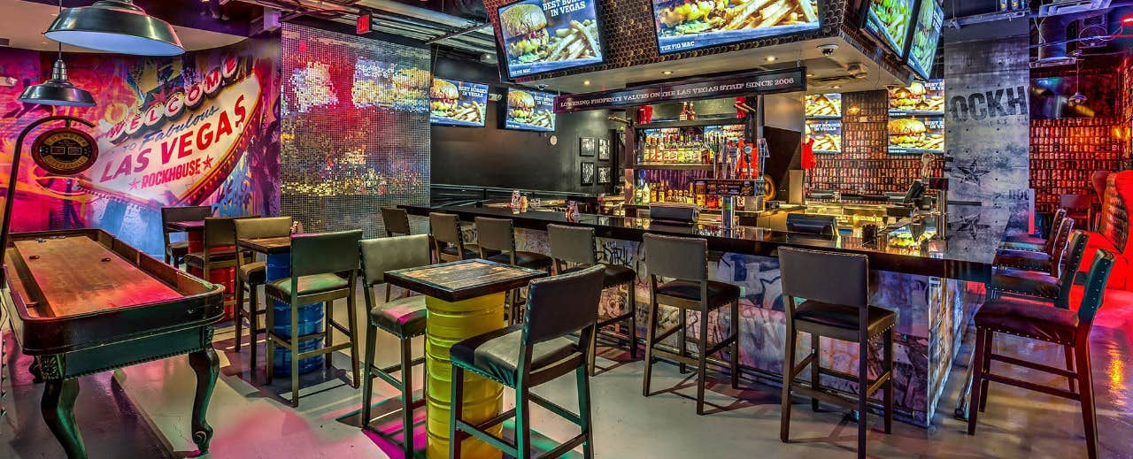 A colorful bar with stools, multiple TV screens, various lights, Las Vegas decor, and a shuffleboard table.