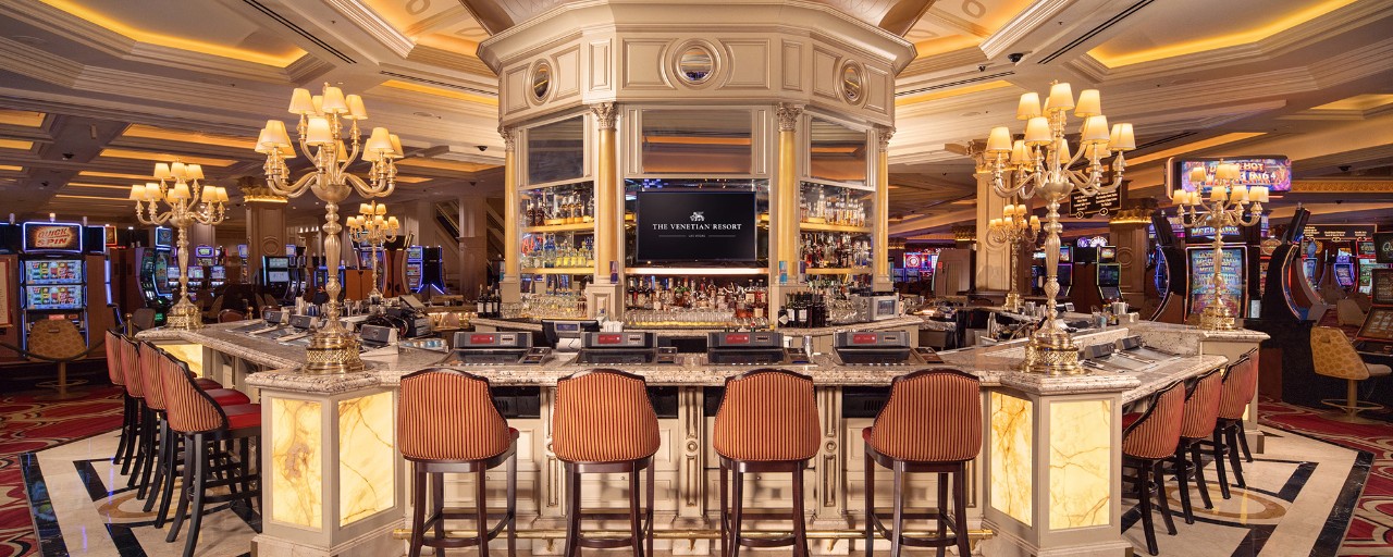 Luxurious casino bar with marble counters, chandeliers, plush bar stools, and slot machines in the background.