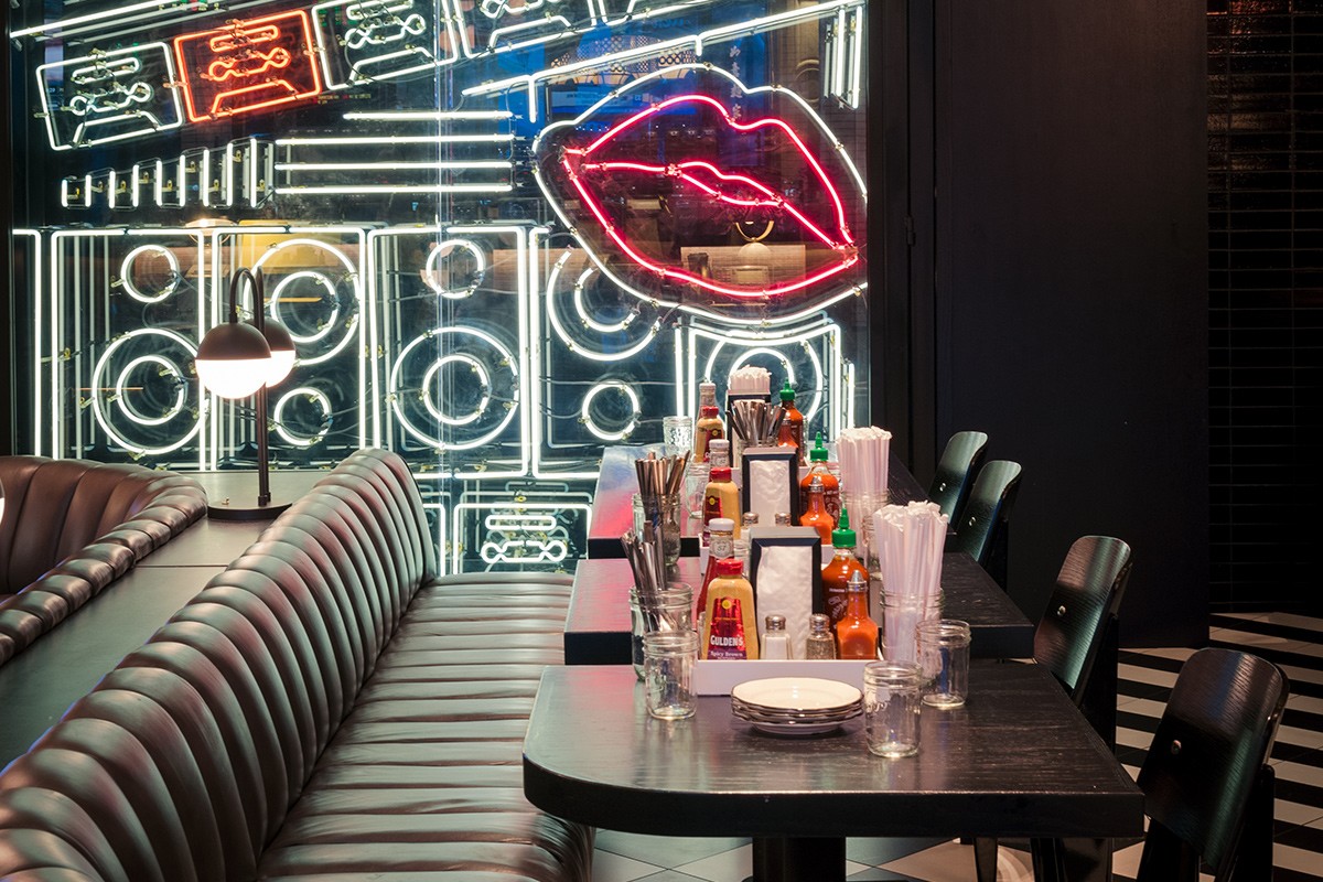 A diner booth with condiments and utensils on the table is set against a neon-lit wall featuring lips and cassette tapes.