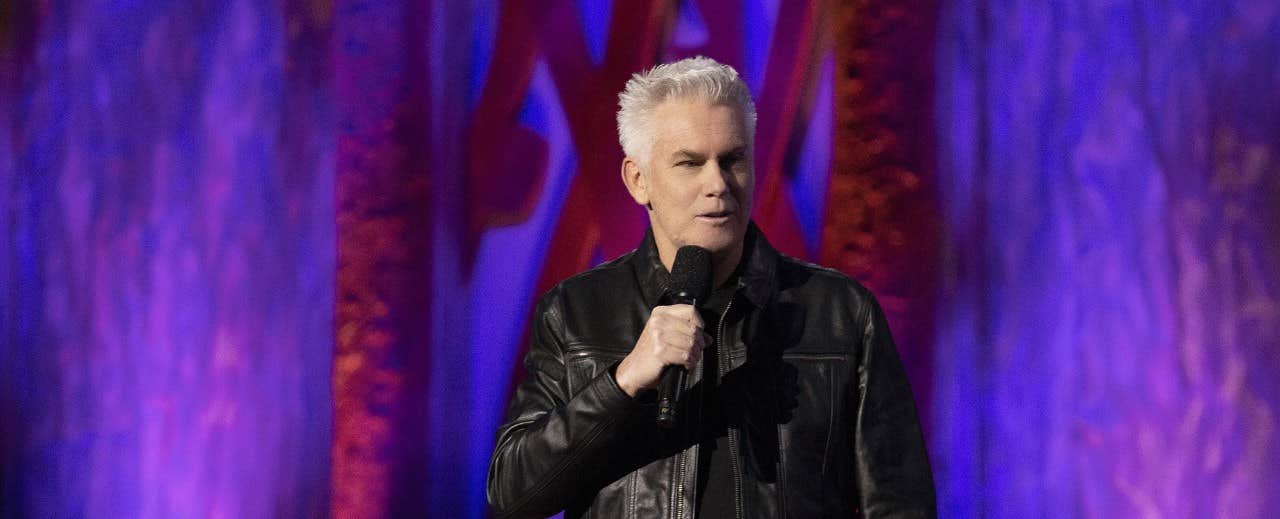 A man with white hair, holding a microphone, performs on stage with purple and red background lights.