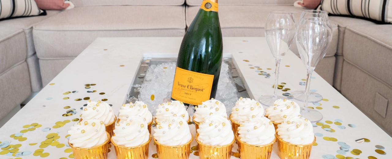 A bottle of champagne surrounded by six cupcakes in gold wrappers and two empty champagne flutes on a confetti-covered table.