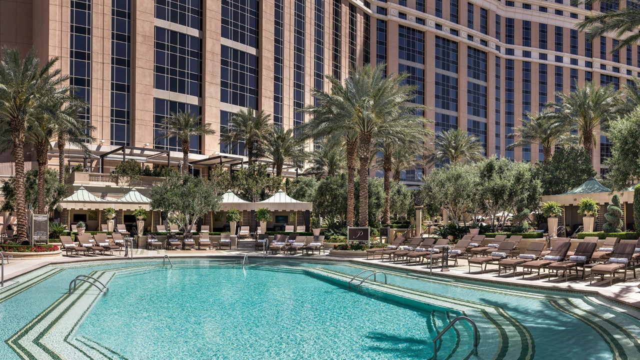 Luxurious hotel pool area with lounge chairs, cabanas, palm trees, and a large building in the background.