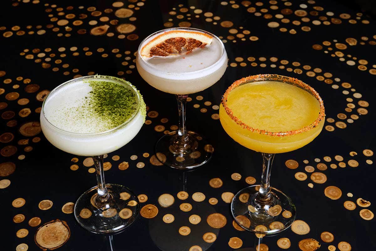 Three cocktails in coupe glasses on a dark table with circular patterns. Each drink is uniquely garnished.