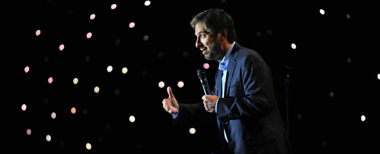 A man in a dark suit holding a microphone and performing stand-up comedy on a stage with a dark, starry background.