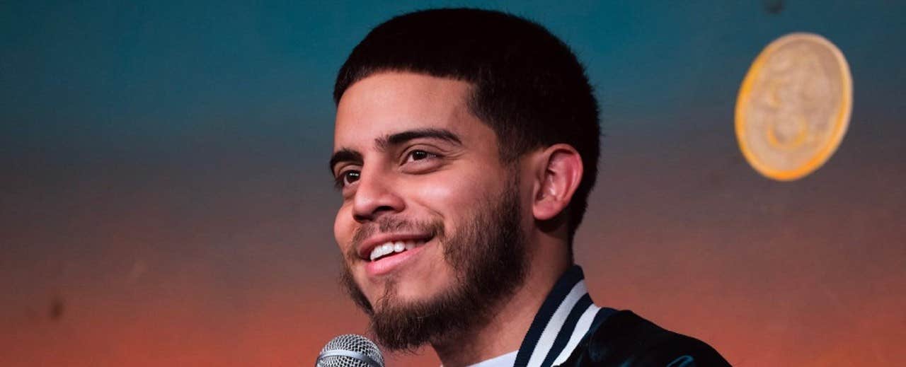 A man with a beard and short hair smiling and holding a microphone against a blurred colorful background.