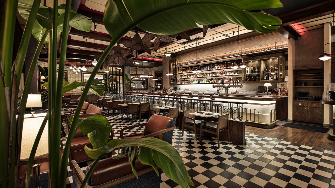 A chic, modern restaurant with checkered flooring, leather seats, a stocked bar, and large green plants in the foreground.