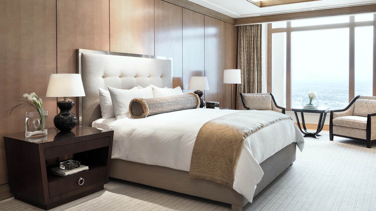 Luxury hotel room with a queen bed, bedside tables, armchairs, and a large window providing a city view.