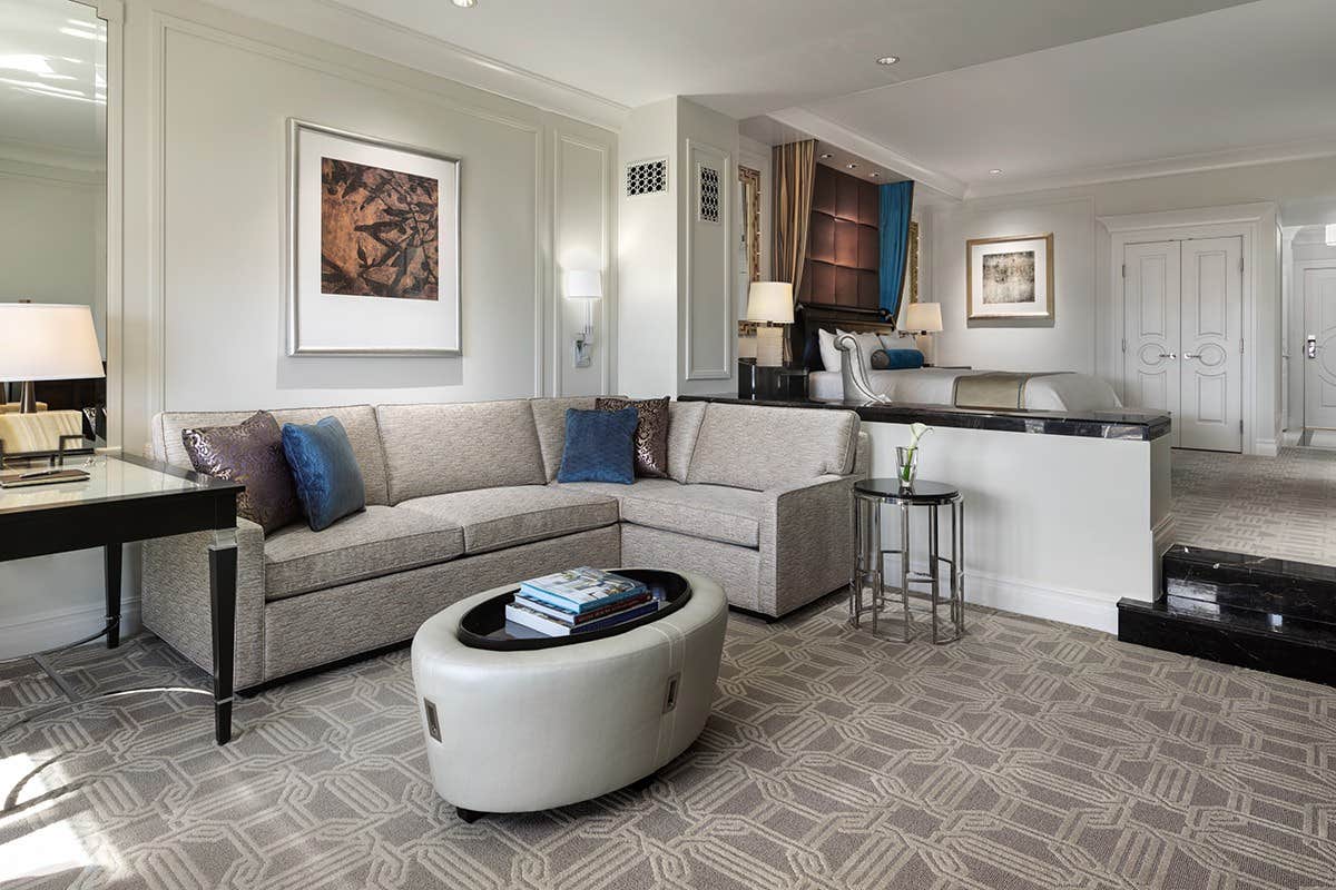 A modern living room with a beige sectional sofa, glass coffee table, wall art, and large windows with light-colored curtains.