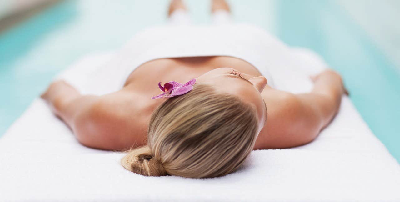 Person lying on their back on a massage table with a pink flower in their hair, covered in a white towel near a pool.