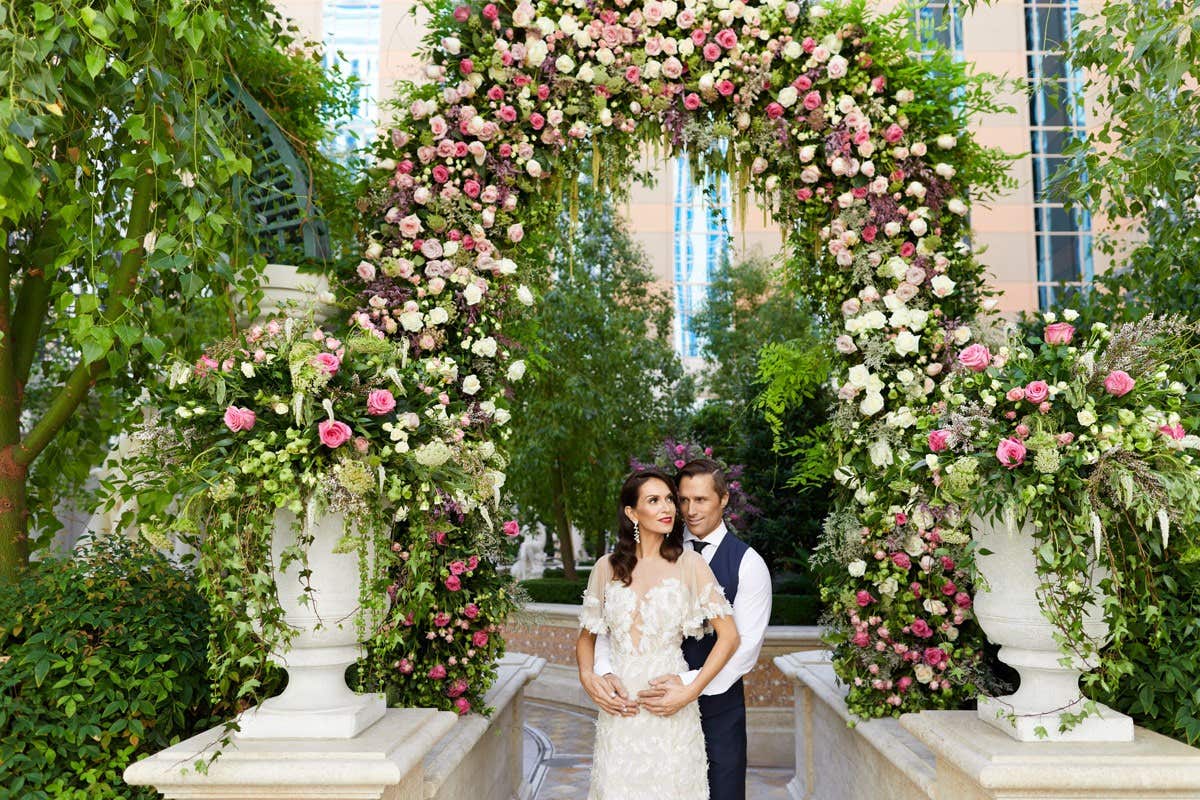 A married couple stands under a floral archway with lush greenery and colorful flowers, in an outdoor garden setting.