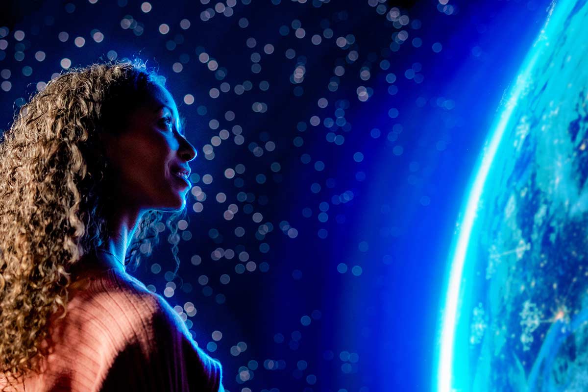 A woman stands in profile, illuminated by glowing blue light from a large, detailed image of Earth in space.