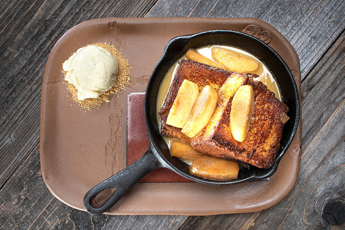 French toast topped with caramelized apples in a skillet, served with a scoop of vanilla ice cream on a brown tray.