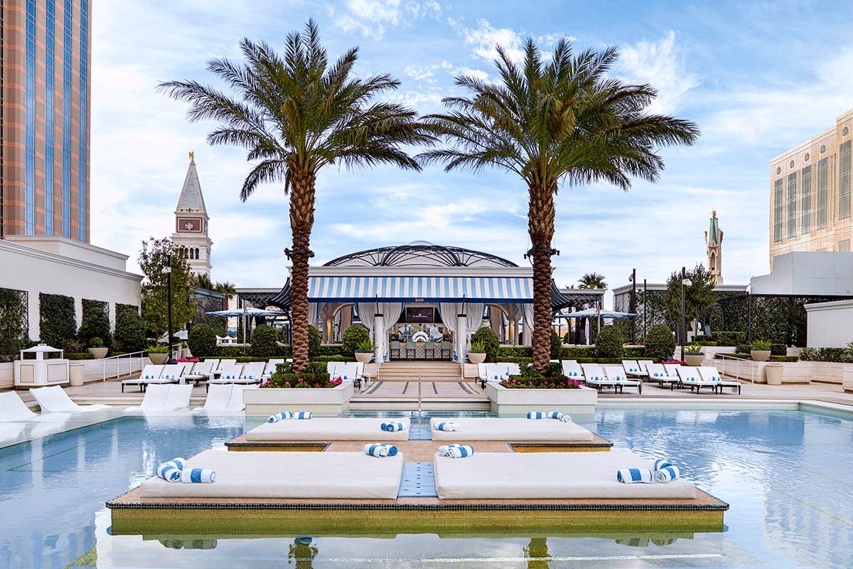 Luxury poolside area with sun loungers, cabanas, palm trees, and a beautiful sky in the background.