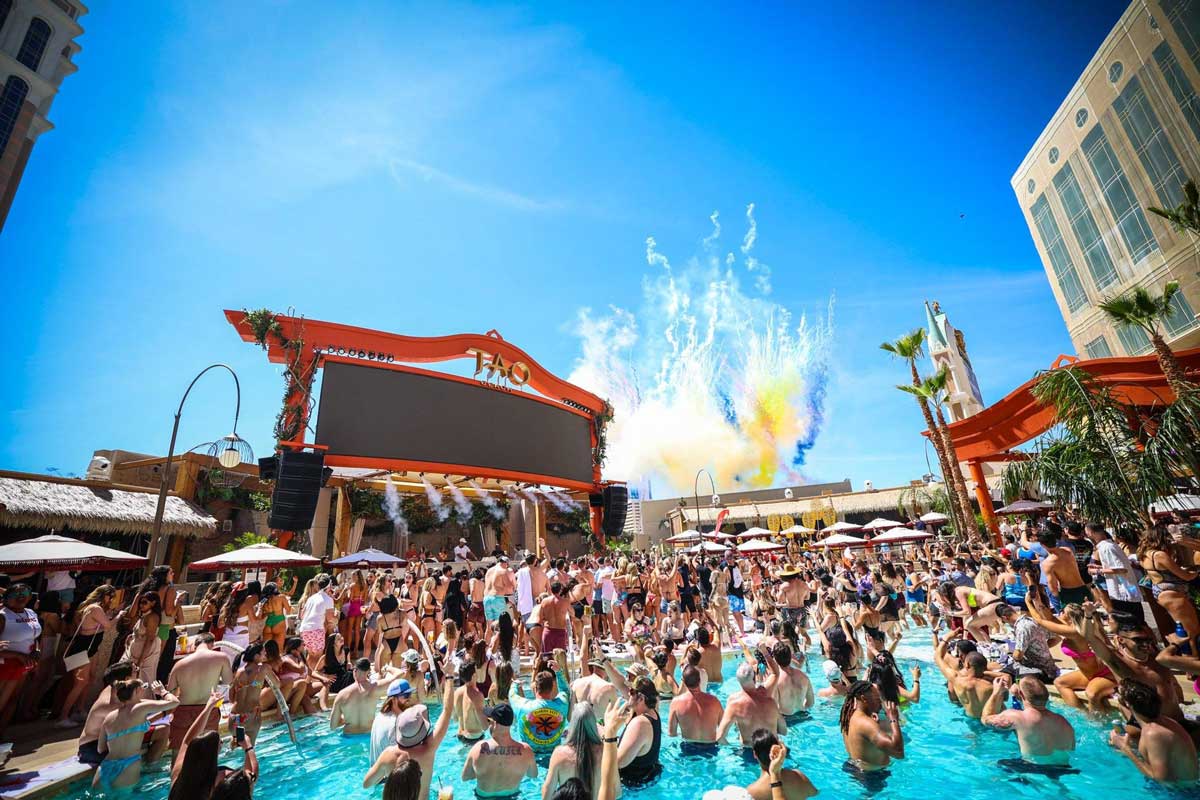 A lively daytime pool party with a crowded pool, a large screen, and colorful fireworks in a sunny outdoor venue.