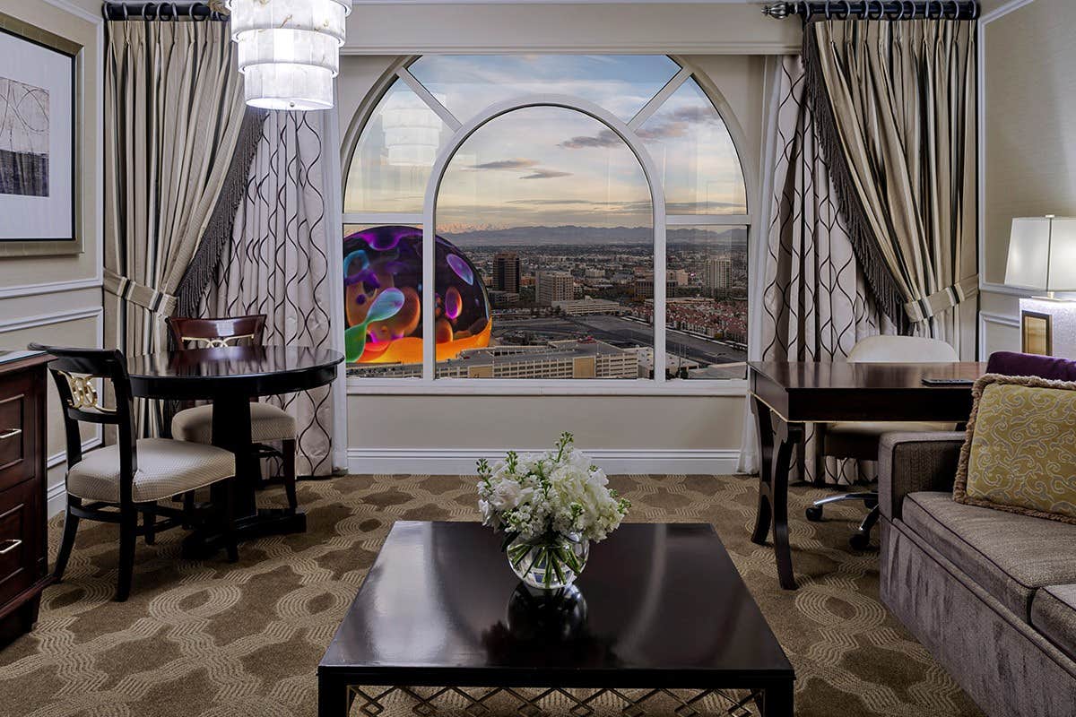 A luxurious hotel room with a large window showcasing a cityscape and colorful LED display, furnished with elegant decor.