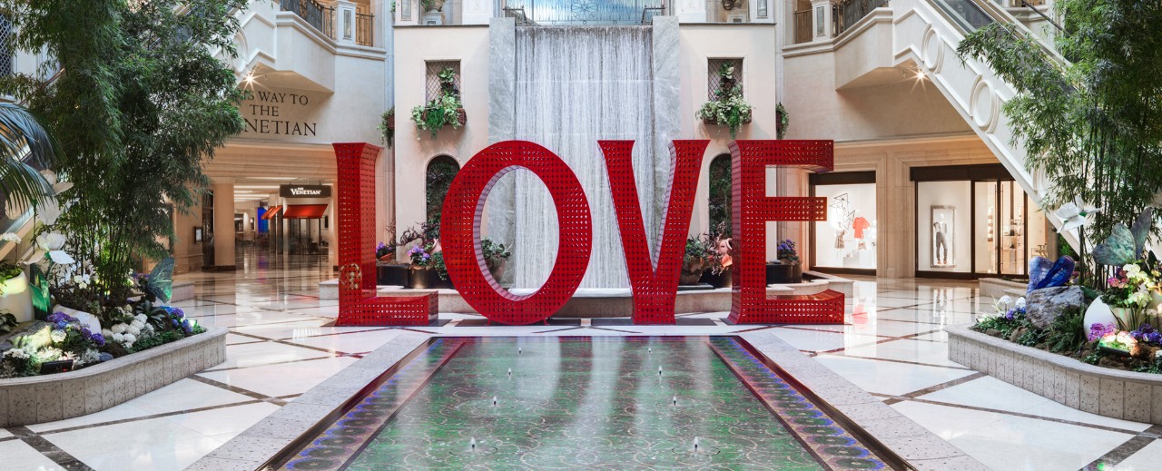 Large red "LOVE" sculpture in an elegant atrium with greenery, shops, and a grand staircase.