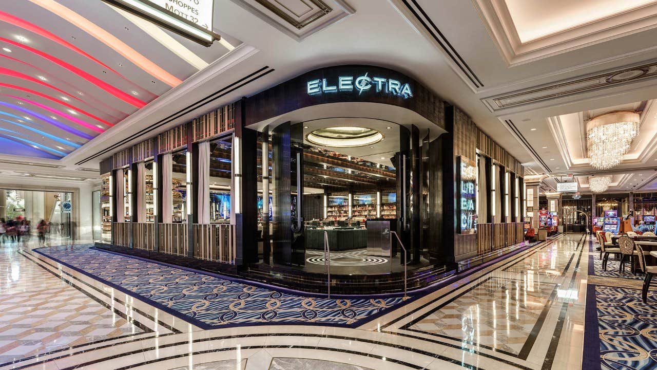 Elegant bar named "Electra" with modern decor in a lavish, brightly lit hallway with a patterned floor and ornate chandeliers.