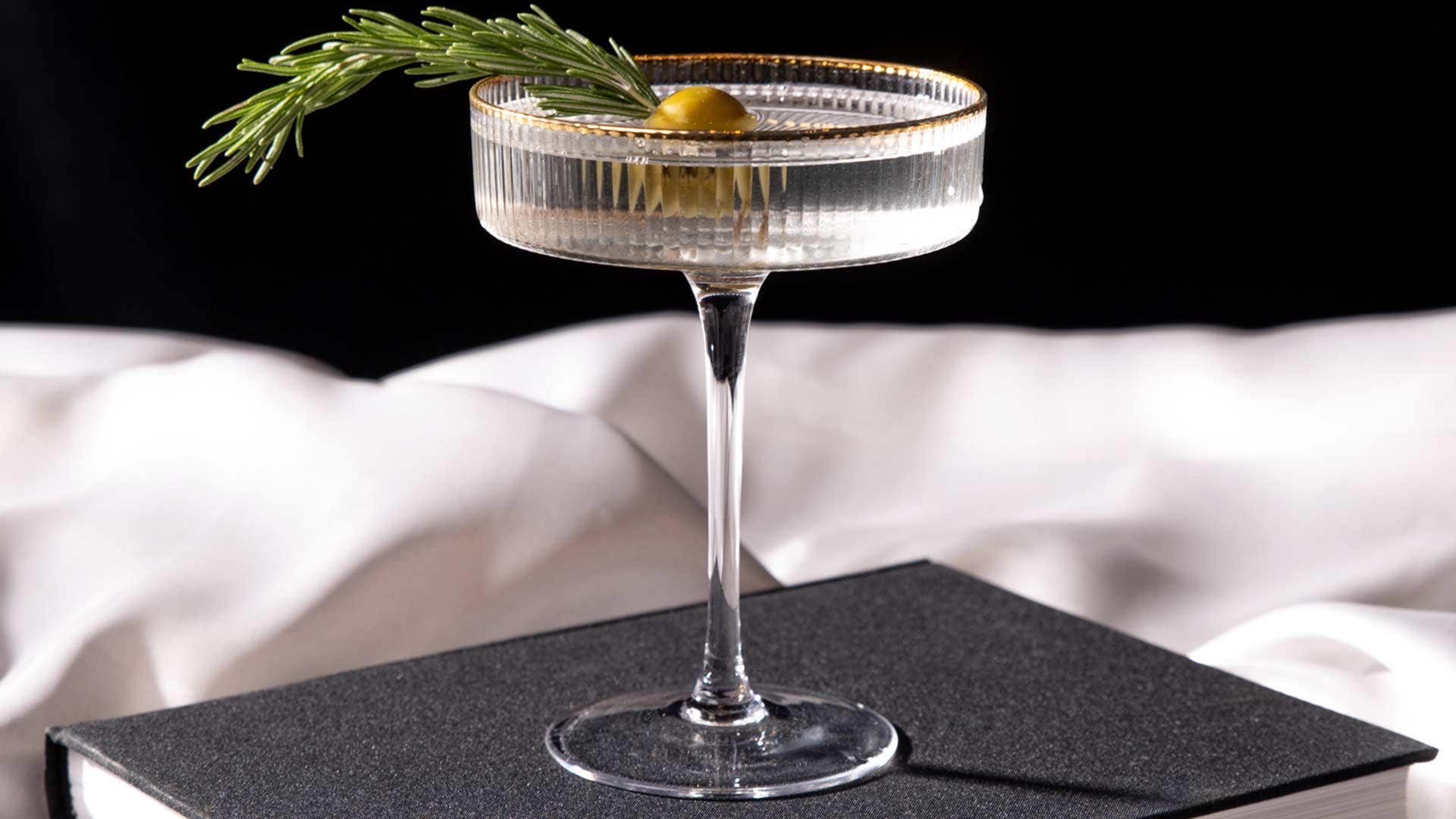 A cocktail garnished with an olive and rosemary sprig, served in a glass, placed on a dark book with a white cloth backdrop.