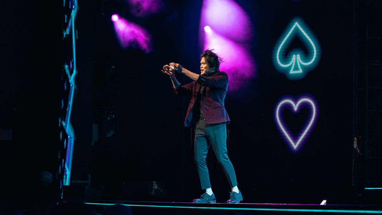 Shin Lim on stage with purple and blue lighting, holding an object with neon spade and heart symbols in the background.