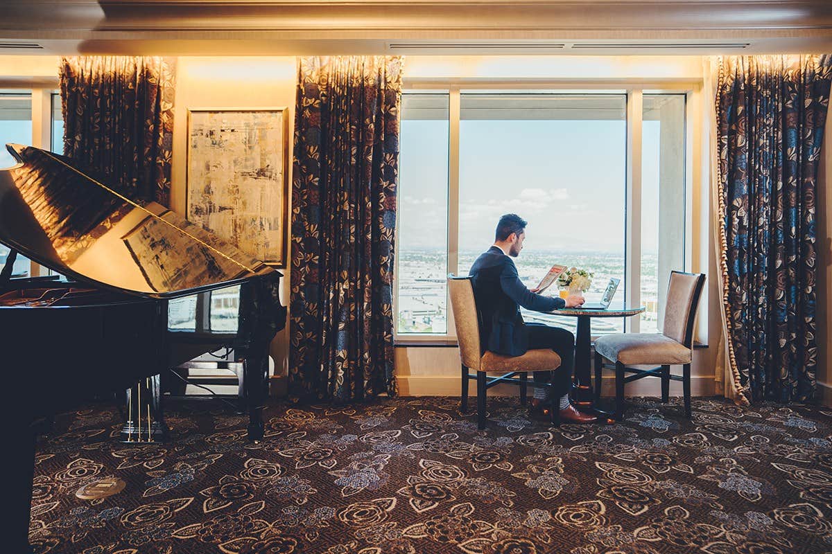 A man in a suit sits at a table with a view, using a laptop in a luxurious room with a grand piano and large windows.