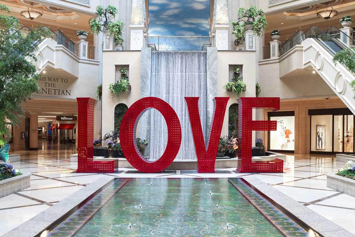 Large red "LOVE" sculpture in front of a water feature inside a shopping mall with ornate architecture and plants.