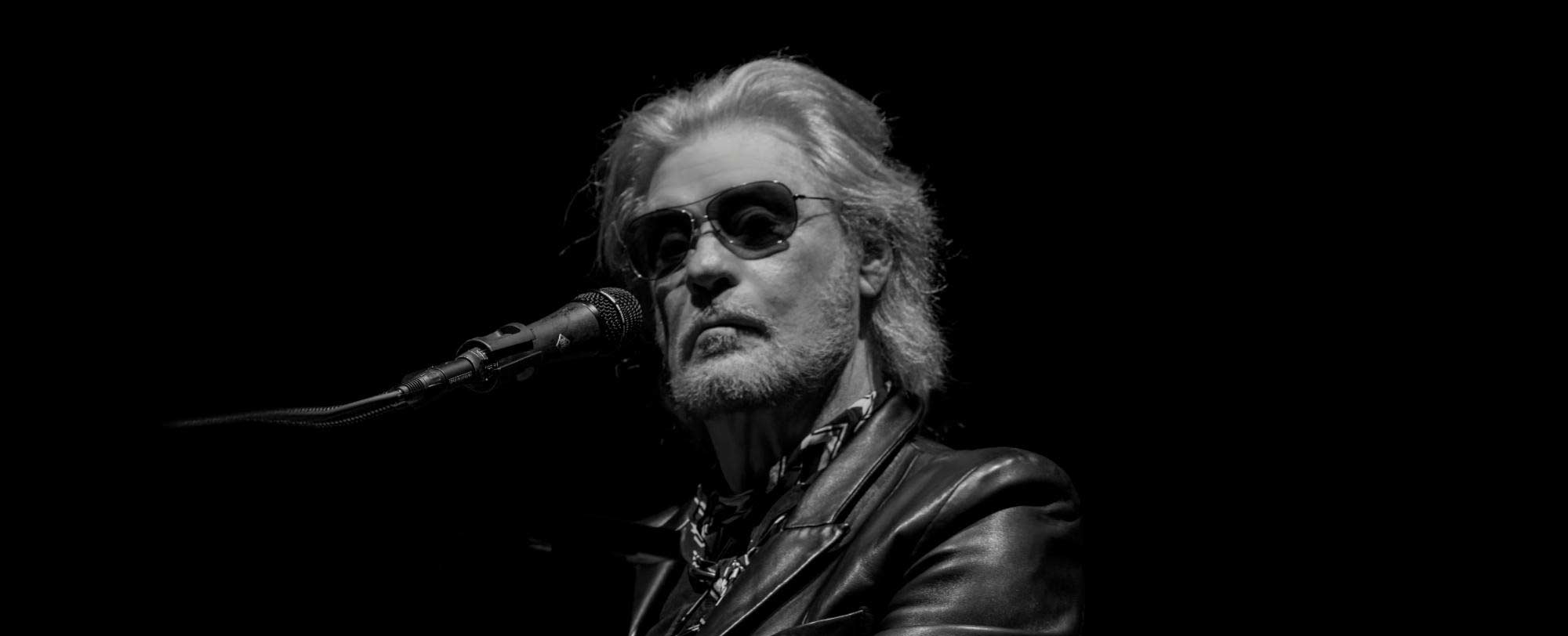 Black and white image of a man with sunglasses singing into a microphone on stage.