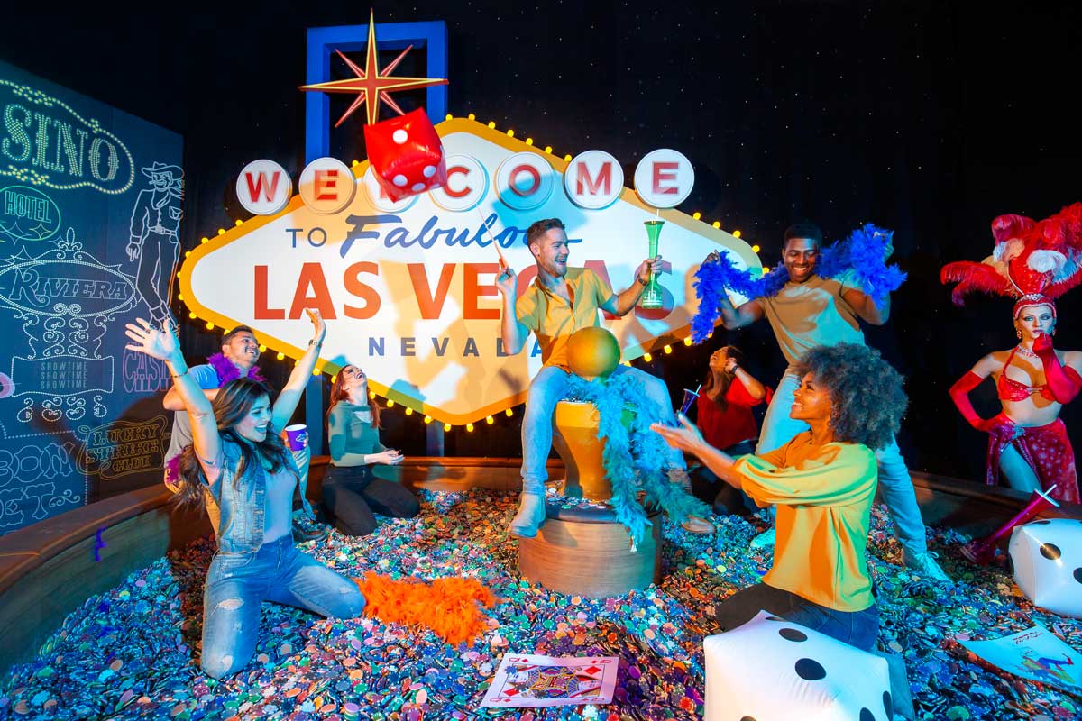 A group poses energetically in front of a bright "Welcome to Fabulous Las Vegas" sign, with confetti scattered on the ground.